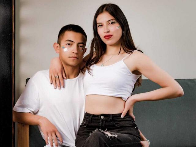 CrimsonSecretts with small tits and a slim ass, wearing a white crop top and ripped jeans, sits with arm around her short-haired boyfriend in a white tee.