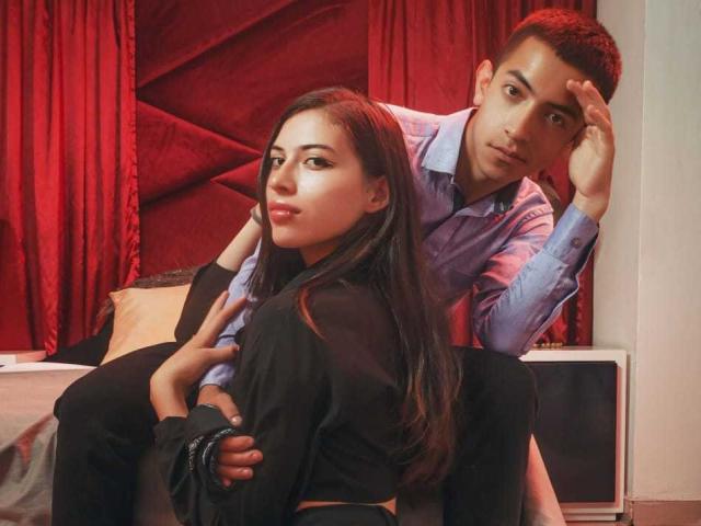 CrimsonSecretts, with long dark hair, sits on a bed wearing black clothes, while a guy in a blue shirt leans over her, hand on her ass, looking confused.