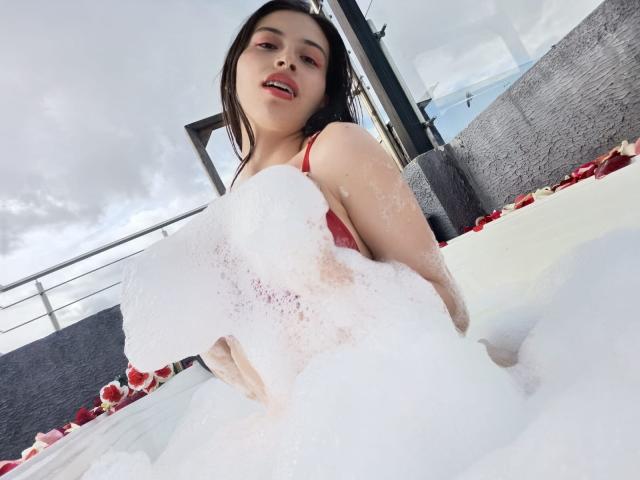 CrimsonSecretts with foamy white bubbles covering her red bra-covered tits, sitting in a bubbly tub, red rose petals around, looking at the camera with a slight smile.