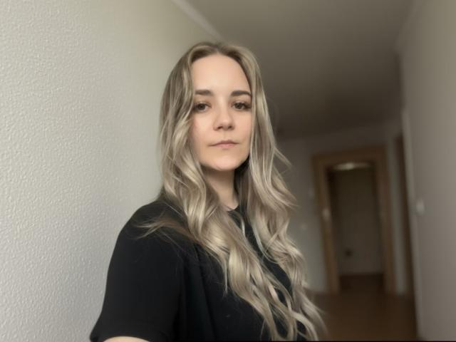 CandiCage has long, wavy blonde hair, a black shirt, and a calm expression. Her small tits are slightly visible under the shirt. She stands in a hallway.