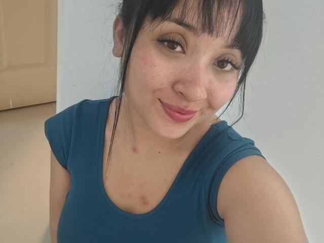 LunaBellaHotty has a medium build with fair skin and a few small red bumps on her chest. She has dark, straight hair with bangs, and is smiling at the camera. She wears a teal, short-sleeve shirt. Her brown eyes are slightly upturned, and her pink lipstick contrasts with her natural look. The image is a close-up selfie with soft lighting. Her expression is gentle and relaxed.