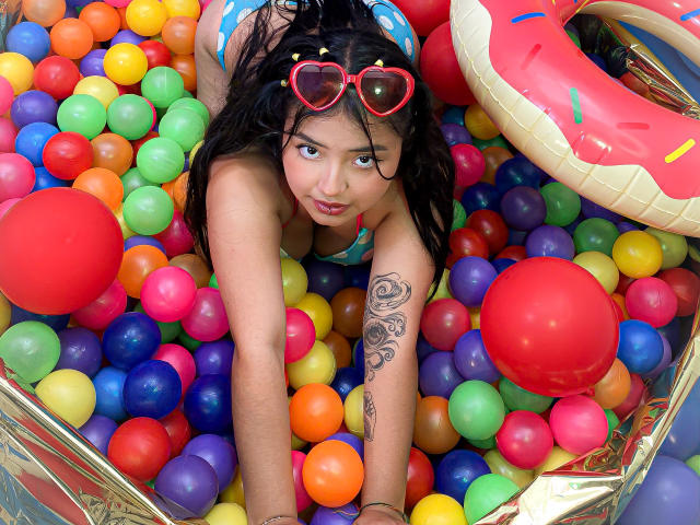 SofiDollg with heart-shaped red sunglasses, in a colorful ball pit, leaning forward, showing tattooed arm, wearing blue bikini, looking up with playful expression, red balloon in foreground.
