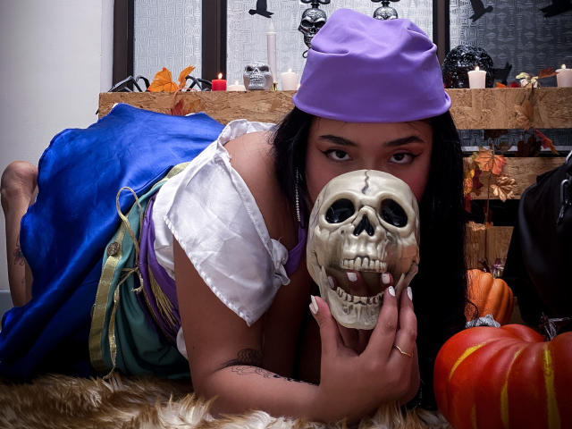 SofiDollg with dark hair, wearing a purple hat, white shirt, blue dress, holding a skull, lying on a furry rug, with a pumpkin and candles in the background.