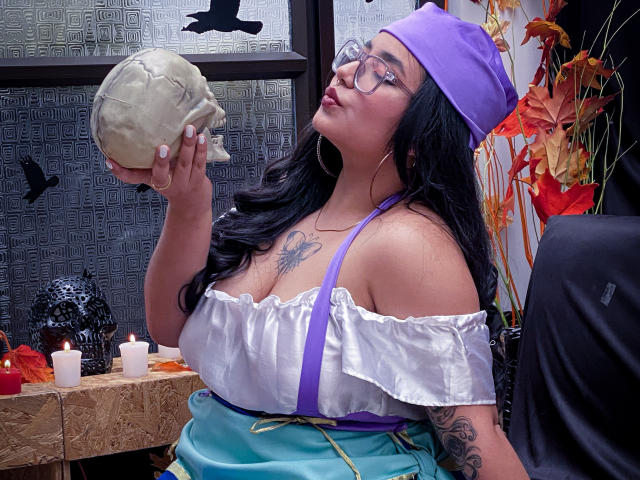 SofiDollg with big boobs, wearing glasses, purple hat, white off-shoulder top, green skirt, holding skull, tattoos on chest, in Halloween-themed setting, candles, autumn leaves in background.
