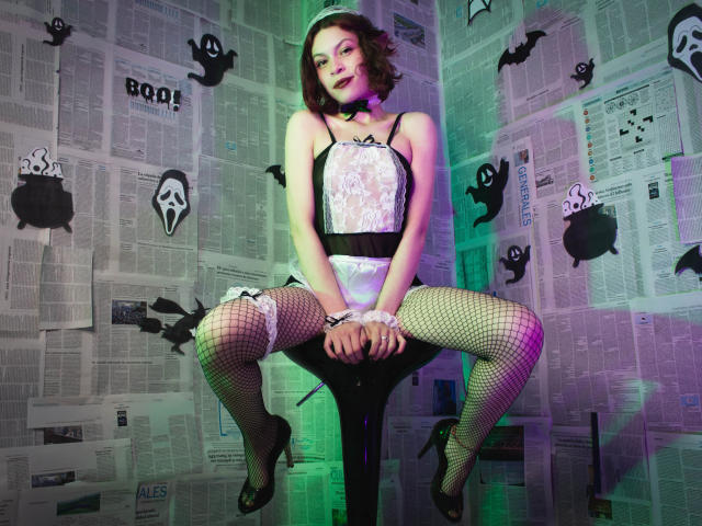 SophieBlake in a white lace corset, black fishnet stockings, and heels, sitting with legs spread, wearing a choker, short brown hair, green and purple lighting.