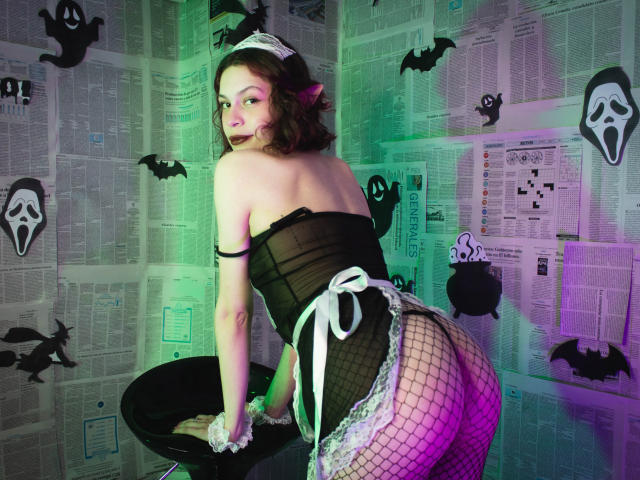 SophieBlake in a black corset and fishnet stockings, bent over a stool, with her ass up, wearing a white lace maid headpiece, and a sexy, seductive face.
