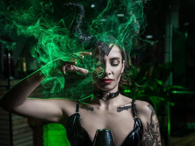 AmberGreyy with green smoke, black horns, and a leather top, showing off tattoos and a choker, her eyes closed, and her hand making a peace sign. Boobs visible.