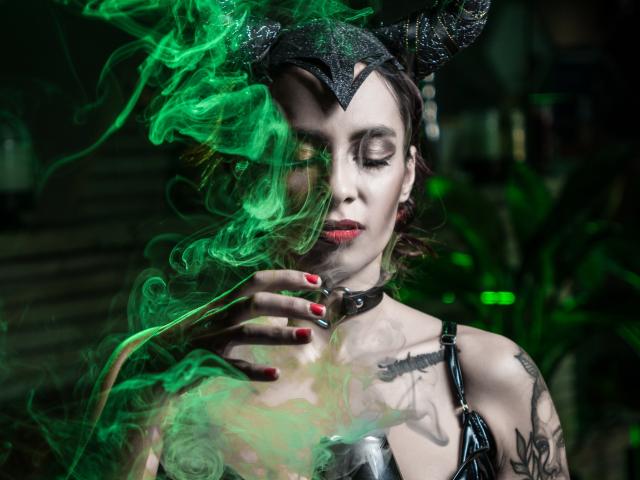 AmberGreyy with green smoke, black headpiece, red nails, choker, tattoos, and latex top, eyes closed, hand near mouth, smoking, dark makeup, goth vibe.