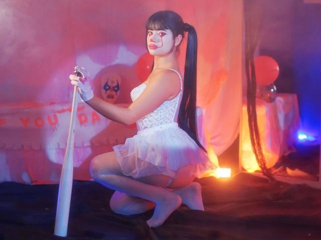 SofiMontoya, with long black pigtails, kneels in white lace lingerie and sheer tulle, holding a silver microphone, wearing white gloves, her ass prominently displayed, in a dimly lit room.