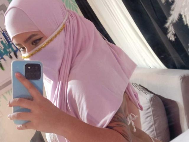 DaliilaAli in a pink hijab, showing her sideboob and tattooed thigh, taking a mirror selfie with a blue phone, her face partially visible with a gold trim.