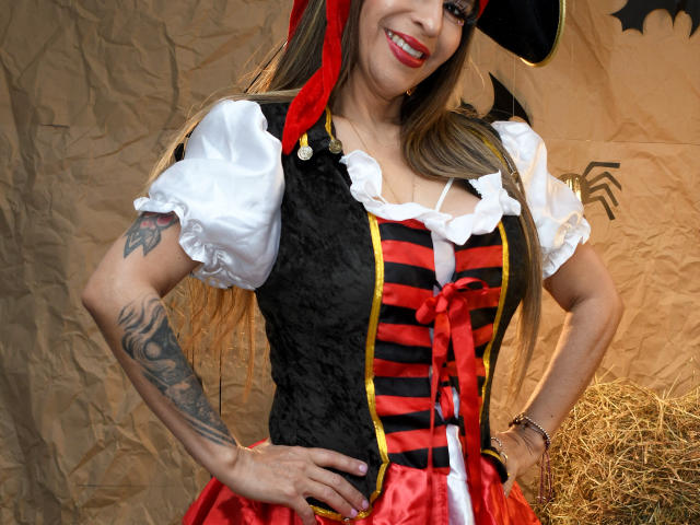 VictoriaJhonnes stands with hands on hips, wearing a pirate costume. She has long brown hair, a red headscarf, and a black pirate hat. Her white puffed sleeves contrast with a black, red, and yellow striped vest, and a red skirt. She has a large tattoo on her left arm and is smiling with red lipstick. The background is crumpled brown paper with a hay bale on the right. The image is a photograph.