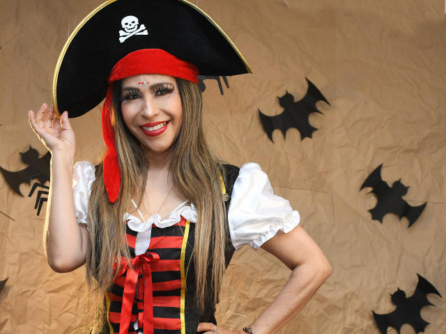 VictoriaJhonnes in pirate costume with black tricorne, red headband, white puffy shirt, red-black striped vest, smiling, long blonde hair, red lipstick, raised hand, brown background with bat shapes.