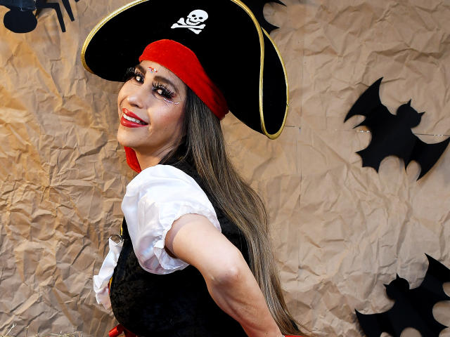 VictoriaJhonnes wears a black pirate hat with a white skull emblem, red headband, and long brown hair. She's smiling with red lipstick, white puffed-sleeve shirt, and black lace vest. Her right arm extends towards the camera. The background has crumpled brown paper with black bat cutouts. Her makeup is bold, with thick eyeliner and long lashes. She has a small forehead tattoo. The image is a selfie.