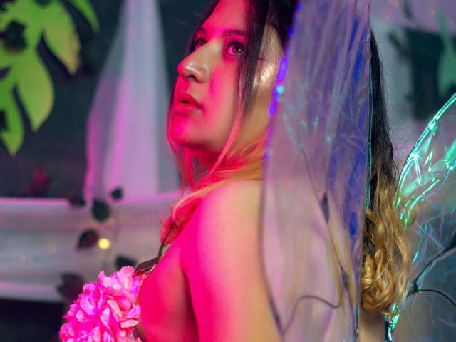 AmaliaRosse with fairy wings, pink flower in hand, neon red and blue light on face, looking over shoulder, wavy blonde hair, slightly parted lips, semi-nude, soft focus.
