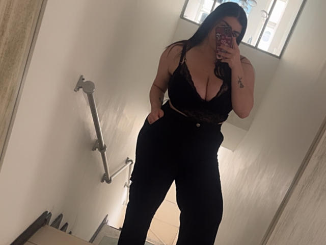 VenusxMusa with big tits in a black lace top and black pants, standing in a bathroom, holding a phone with a red case, covering her face.