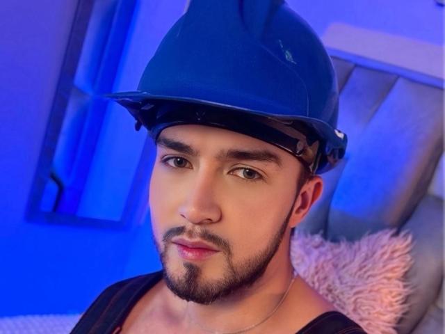 DaltonWhite, com um capacete azul, olhos castanhos e barba, vestindo uma camisa preta, tem um leve sorriso e um travesseiro rosa e felpudo ao fundo..