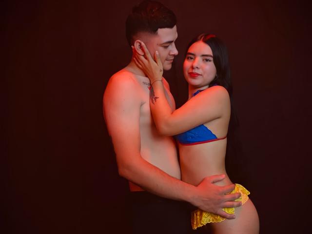 Laura e Kevin, com cabelos castanhos curtos, sem camisa, segurando uma mulher de cabelos longos e pretos, com um sutiã azul e calcinha de renda amarela, em um fundo escuro, com as mãos nos rostos e quadris uma da outra..