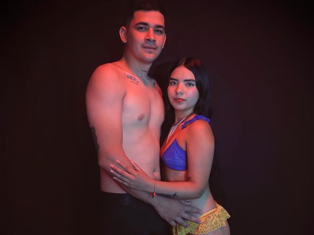 LauraAndKevin, a tall, shirtless man with short dark hair and a tattoo on his right shoulder, stands with a short-haired woman in a blue lace bra and yellow patterned pants. He has a slight smile, and she looks at the camera with a neutral expression. He has his left arm around her, and she rests her right hand on his chest. The man's skin is light, and the woman's skin is slightly darker. The lighting is red, creating a warm, intimate atmosphere.