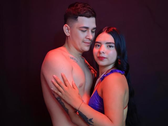 Laura e Kevin, sem camisa, com tatuagens, e Kevin, vestindo um vestido roxo, em pé, próximos, de braços entrelaçados, iluminação vermelha e azul, sorrisos suaves, fundo preto. A mão de Kevin no braço de Laura e Kevin..
