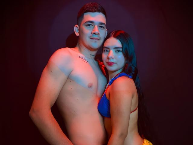 LauraAndKevin, a shirtless man with short brown hair and a tattoo on his left pectoral, stands next to a woman with long black hair wearing a blue bra. Both have light skin and are positioned in the center of the image with red and blue lighting. The man's right arm is around the woman's waist. The woman's left arm is around his back. The man has a slight smile, and the woman looks directly at the camera with red lipstick. The image has a dark, shadowy background.