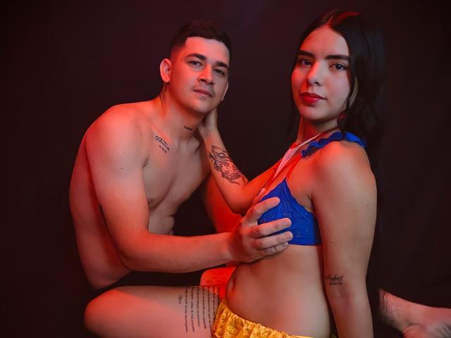 LauraAndKevin, a young man and woman, sit closely in a dimly lit setting. The man, shirtless with tattoos on his chest and arm, holds the woman's blue bra-covered breast with his right hand. He has short, dark hair and a slight smile. The woman, with long dark hair and a confident expression, wears a blue bra and yellow shorts. Her left arm has a tattoo. The image uses red and black lighting, creating a dramatic, intimate atmosphere.