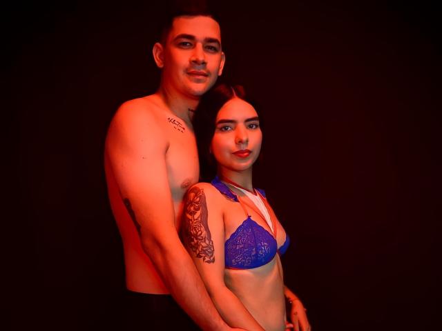 LauraAndKevin, ohne Hemd und mit einem Tattoo auf seinem linken Arm, steht hinter LauraAndKevin, die ein blaues Spitzen-BH trägt, beide beleuchtet mit rotem Licht, ihre Brüste sind sichtbar, sein Penis wird nicht gezeigt..