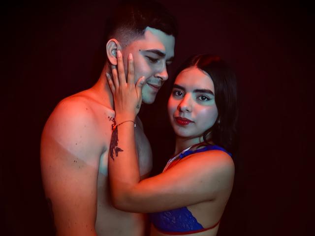 LauraAndKevin, een charmante vrouw met rode lippenstift, raakt de wang aan van een naakte man met een tatoeage, beiden staan in een kamer met rood licht en dragen blauwe lingerie..