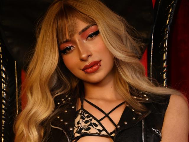 OasisFairy has blonde wavy hair, red eyeliner, and glossy orange lips. She wears a black studded leather vest over a black lace-up top, showing small tits.