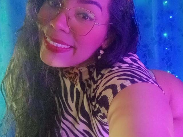 FetishLoverAna with long black hair, wearing glasses and a zebra-print top, smiling at the camera, showing her round ass. Blue and purple lights in the background.