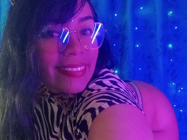 FetishLoverAna with glasses, zebra print shirt, and pinkish-blue lighting, smiling at camera, showing her curvy ass in tight clothes, dark hair, red lipstick, and glowing eyes.
