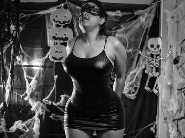 Black and white photo of MorganMoretz in a tight, shiny leather mini-dress, wearing a masquerade mask, arms behind back, showing large, perky tits, and a curvy, sexy ass.