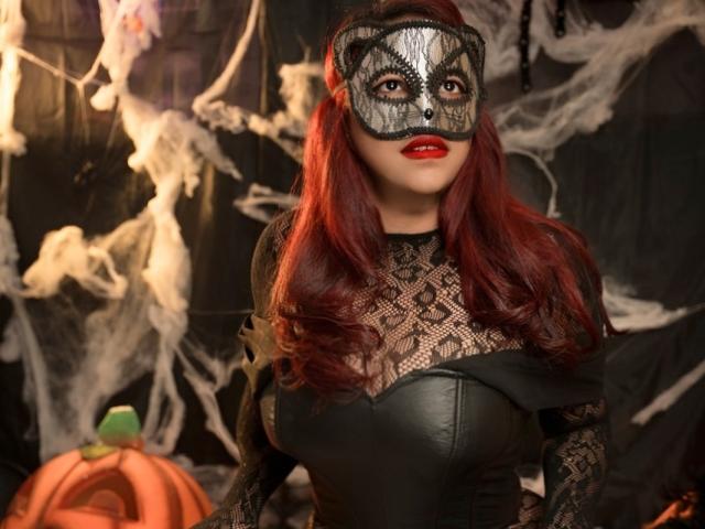 MorganMoretz with red hair, black lace mask, and red lipstick, wearing a black lace dress, showing her big tits, standing in front of a pumpkin.
