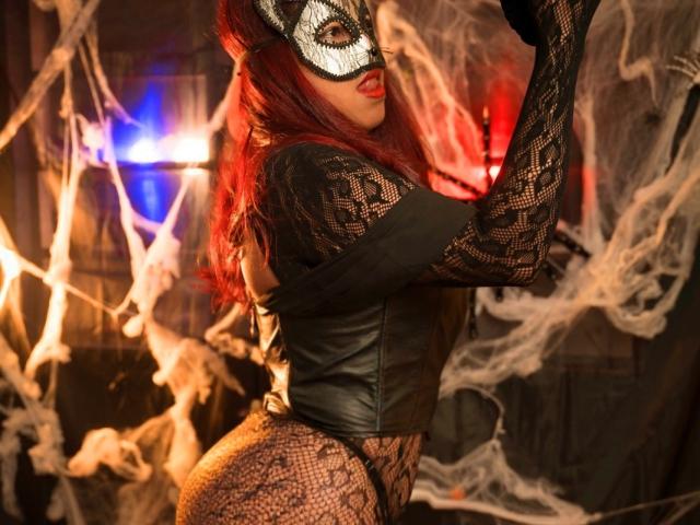 MorganMoretz with red hair, black lace mask, leather crop top, and fishnet bodysuit, showing her ass, in a dark, spooky, smoky setting.