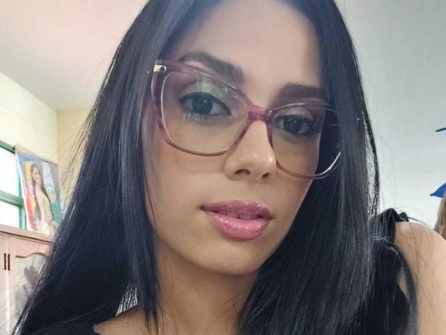 AnaisHilton with long black hair, wearing pink glasses and shiny pink lipstick, looking into the camera with a slight pout. Her tits are slightly visible under a black top.