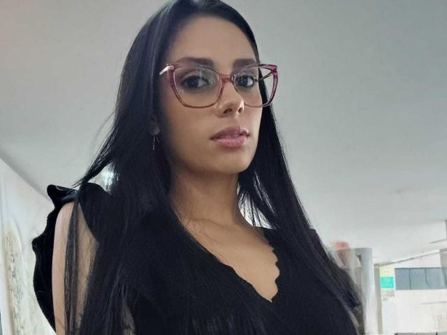 AnaisHilton with long black hair, wearing red glasses and a black top, showing her tits and a slight smile, looking at the camera in a casual photo.