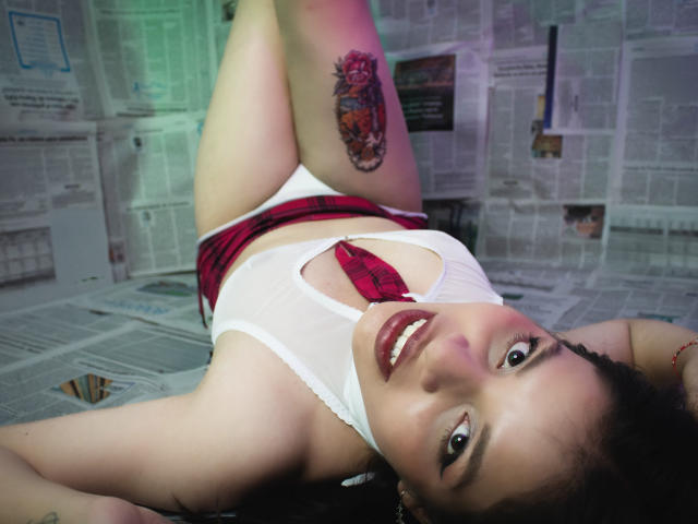 AllanaMicheles, allongée sur le dos, porte un débardeur blanc, une mini-jupe à carreaux rouges et des sous-vêtements blancs, et on peut voir un tatouage sur sa cuisse droite. Elle sourit à l'appareil photo..