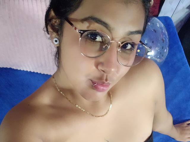Emiily Johamson, con gafas, labios fruncidos y un collar dorado, hombros desnudos y senos pequeños, con aretes grandes, mirando hacia arriba, sobre superficies azules y rosas..