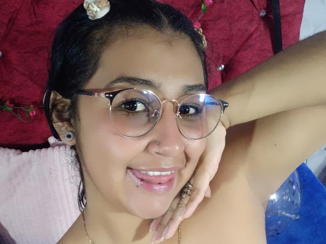 Emiily Johamson, avec des lunettes, la langue tirée, peau brune, petit anneau de nez, cheveux noirs avec des coquillages, boucles d'oreilles, bras levé, souriante, lèvres roses, lèvres visibles..