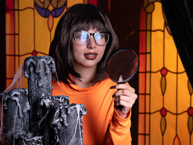 CamilleRios with dark brown bob, glasses, orange shirt, holding mirror, examining black, waxen candles dripping with white goo, looking slightly annoyed.