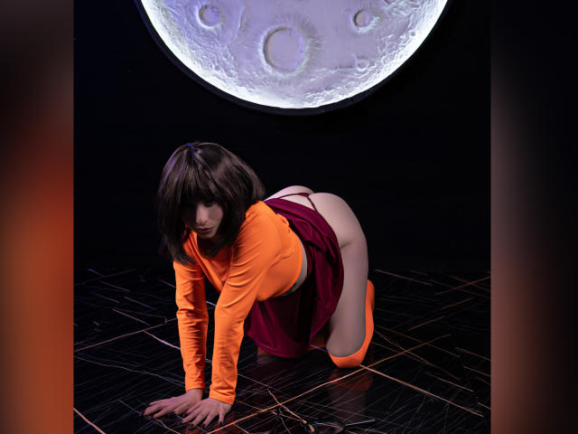 CamilleRios is on all fours, wearing an orange top and purple skirt, exposing her bare ass. She has brown hair, and a large moon is in the background.
