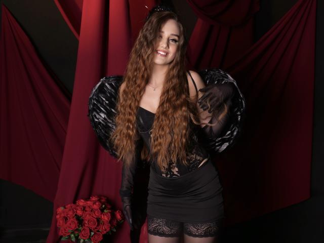 SabrinaCandi with long wavy brown hair, black lace top, black mini skirt, and black feathered wings, smiling, wearing black gloves, standing in front of red curtains with roses.