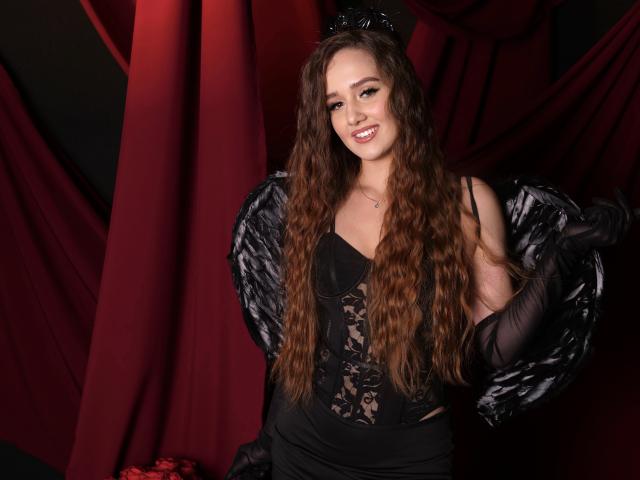 SabrinaCandi con lunghi capelli castani ondulati, un abito nero in pizzo, ali nere piumate, guanti neri e un copricapo nero, sorridente, che mostra il seno, con una tenda rossa sullo sfondo..
