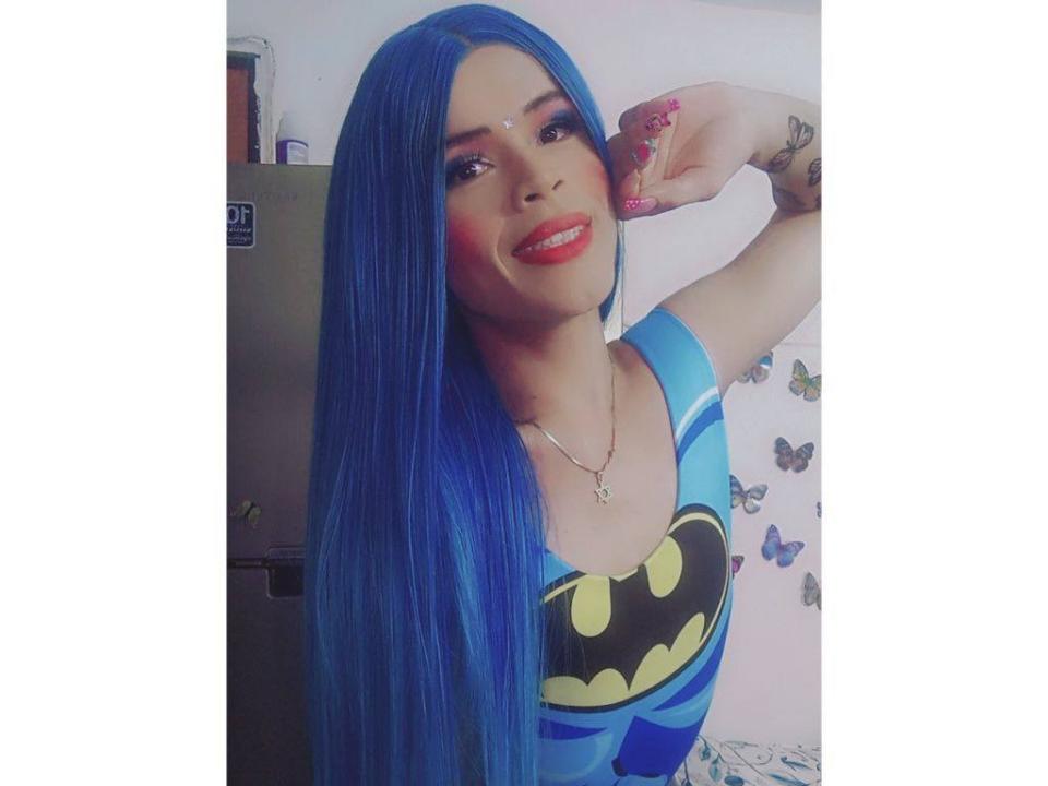 NatalyBusny, avec de longs cheveux bleus, du rouge à lèvres rouge, et un t-shirt Batman, souriante, la main près du visage, montrant des ongles roses, un petit tatouage sur le bras, portant un collier..