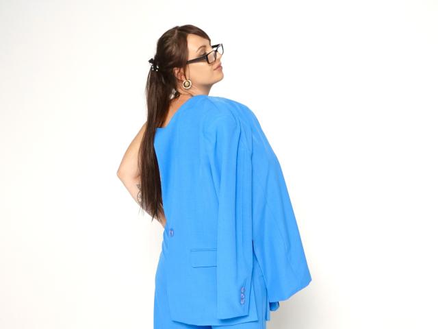 Een vrouw met lang bruin haar, zwarte bril, een blauwe oversized jas en oorringen, kijkt over haar schouder en laat een tatoeage op haar linkerarm zien, op een witte achtergrond..