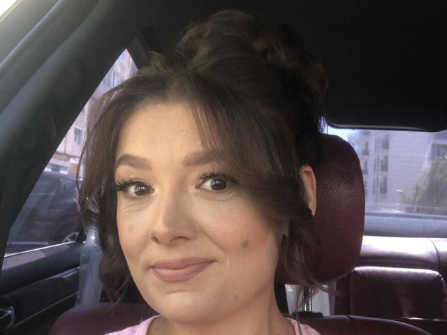 Close-up of MayaHt in a car. She has light brown skin, dark brown hair in a messy bun, and subtle makeup. She has small moles on her left cheek and right side of her nose. She wears a pink top. Her eyebrows are dark and slightly arched. She's looking slightly to the right with a neutral expression. The car's interior features red leather headrests and a window on the left. Natural light illuminates her face.