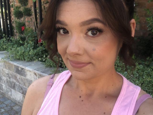 Close-up of MayaHt with medium brown wavy hair, light skin, and brown eyes. She has long, dark eyelashes and a small black mole on her right cheek. MayaHt wears a pink tank top with purple straps, and her makeup includes subtle pink lipstick and minimal eyeliner. She's looking slightly to the left with a soft smile. The background includes green plants and a brick wall. The image is brightly lit with natural light.