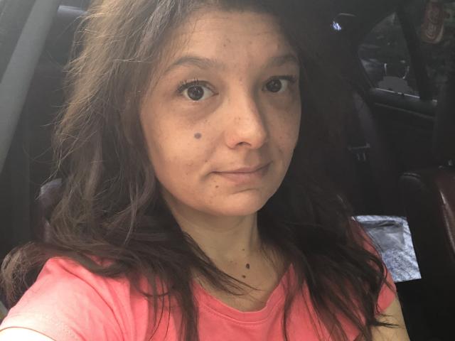 A close-up selfie of MayaHt. She has long, wavy brown hair, light skin, and brown eyes. MayaHt wears a pink shirt and has small dark moles on her right cheek and neck. The photo is taken inside a car, with a dark leather seat and a car door visible in the background. Natural light highlights her face, creating subtle shadows. Her expression is neutral, and her lips are slightly pressed together. The image is clear, with a focus on MayaHt's facial features.