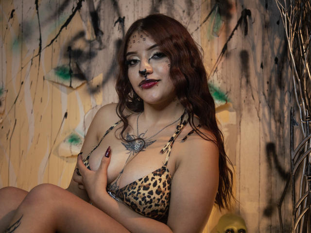 MaylinSanchez with red hair, leopard print bra, holding her big tits, nose ring, tattooed chest, dark lipstick, sitting, background graffiti, right arm tattoo, confident smile, dark nails.