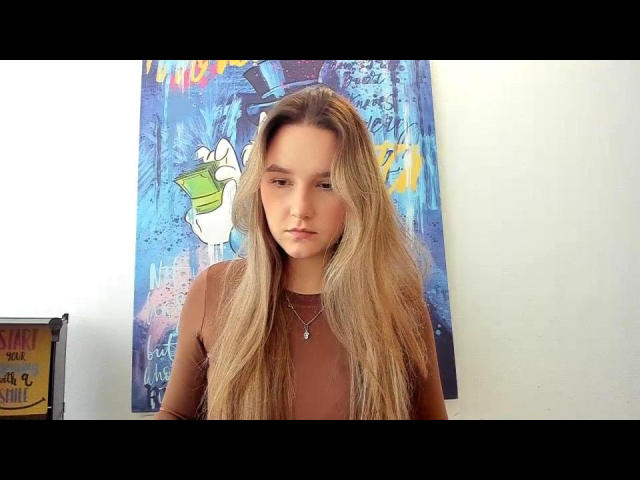 Karpinka with long blonde hair, brown top, and necklace, staring straight ahead, standing in front of colorful graffiti poster, looking serious and cute.