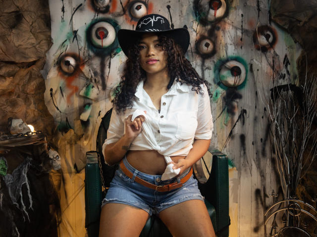 KellyAmbaris with curly hair, wearing a black hat, white tied-up shirt, and denim shorts, sitting on a green chair, showing her tits and a small, visible belly button.